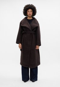 Abrigo marrón texturizado de gran tamaño con un collar ancho, cintura ceñida y mangas largas. Combinado con pantalones de mezclilla oscuros de pierna acampanada.