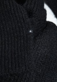 Melna adīta šalle, kas izgatavota no maigas vilnas, ar ribota struktūra un mazu melnu etiķeti ar sudraba zvaigžņu akcentu.