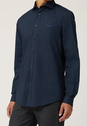 Mężczyzna ubrany w ciemnoniebieską koszulę z długim rękawem zapinaną na guziki, z małym haftowanym logo psa na piersi, w połączeniu z ciemnoszarymi spodniami.