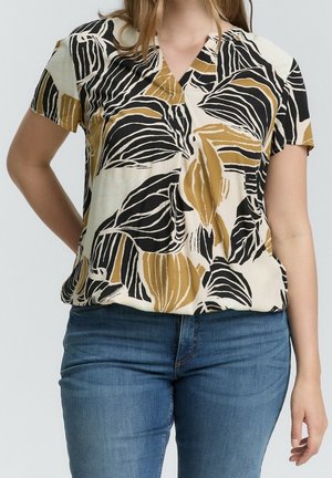 Femme portant un chemisier beige à manches courtes orné de motifs de feuilles noires et marron, associé à un jean bleu, debout avec une main sur la hanche.