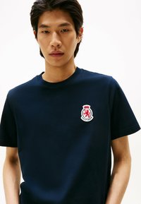 Camiseta de algodón azul marino de manga corta, con cuello redondo y un logotipo bordado circular rojo y blanco en el lado izquierdo del pecho.