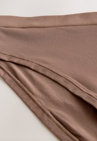 Primer plano de tela lisa de color marrón claro con costuras visibles a lo largo de los bordes doblados, destacando la textura y los detalles de las costuras.