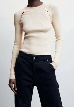 Pull près du corps crème côtelé avec des détails de boutons le long des coutures des épaules, associé à un jean noir taille haute, une main dans la poche.