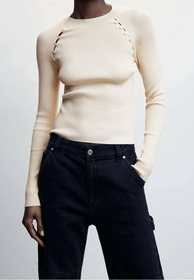 Pull près du corps crème côtelé avec des détails de boutons le long des coutures des épaules, associé à un jean noir taille haute, une main dans la poche.