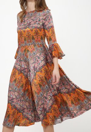 Donna che indossa un vestito a maniche lunghe di media lunghezza con vivaci motivi patchwork arancioni e floreali, muovendo la gonna verso l’esterno.