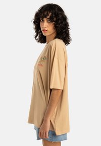 Beige avslappet t-skjorte med korte ermer, rund hals og sideslisser. Har broderte palmer og logo på forsiden.