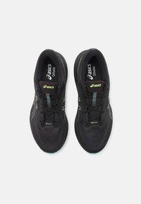 Baskets de sport noires avec une tige en maille, dotées d'accents verts, de lacets noirs et d'une semelle rembourrée. Le logo "ASICS" est visible sur la languette.