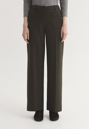 Mujer con pantalones anchos marrones y negros a cuadros y una camiseta gris de manga larga, de pie con zapatos negros sobre fondo blanco.