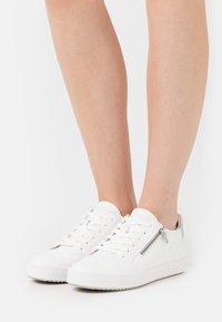 Le sneakers bianche in pelle presentano una punta rotonda, suola piatta, chiusura con lacci e un dettaglio con zip laterale. Il design è minimalista e pulito.