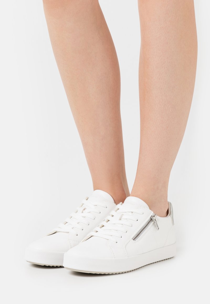 Le sneakers bianche in pelle presentano una punta rotonda, suola piatta, chiusura con lacci e un dettaglio con zip laterale. Il design è minimalista e pulito.
