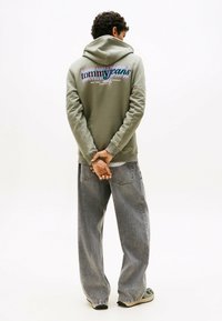 Uomo in piedi con le mani giunte dietro la schiena, indossa un hoody verde di Tommy Jeans e jeans grigi larghi, con sfondo bianco.