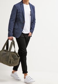 Námořnický blejzr přes bílou košili s černými pruhy, černé kalhoty a bílé tenisky; nese olivově zelenou sportovní tašku.