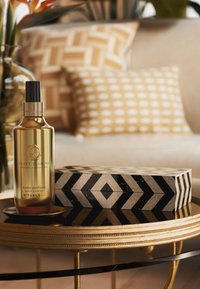Bottiglia spray dorata etichettata "Jasmine Dolce" su un tavolo rotondo con una scatola a motivi in bianco e nero e cuscini decorativi dalle trame morbide.