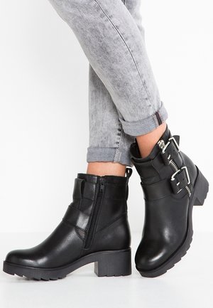 Botas de tobillo de cuero negro grueso con cremallera lateral, detalles de hebilla y un acabado texturizado. El tacón es ancho para mayor estabilidad.