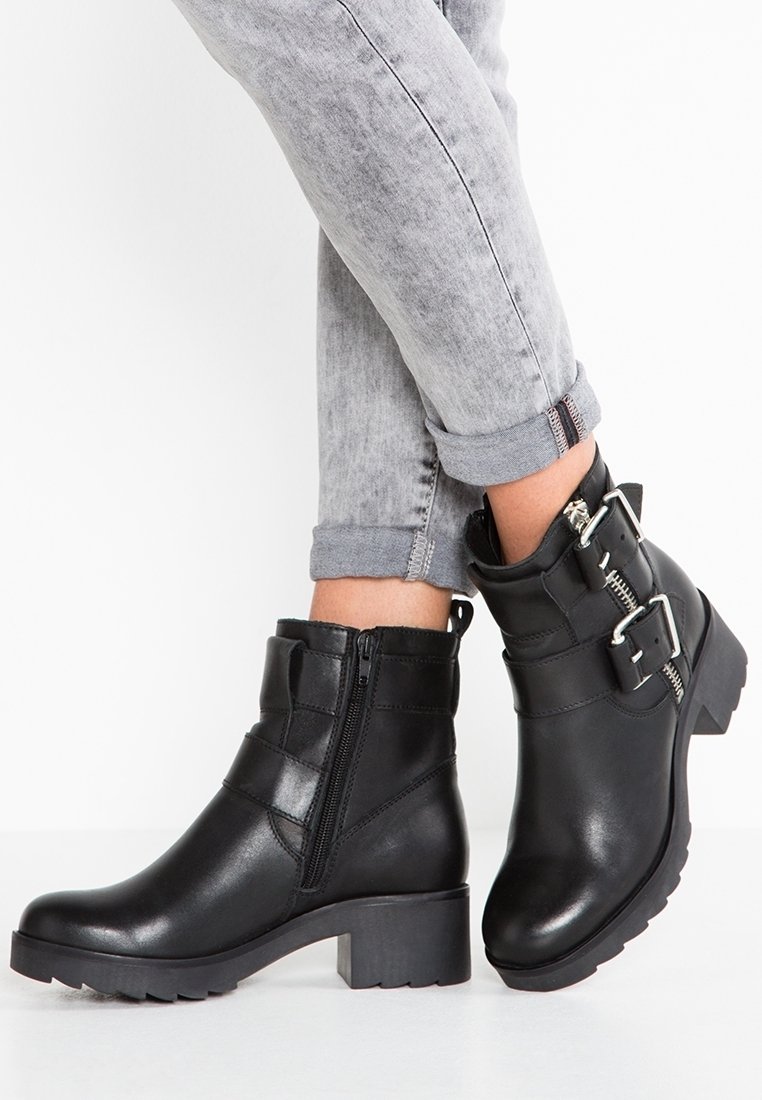 Kraftiga svarta läderankelstövlar med sidodragkedja, spännelement och en texturerad yta. Klacken är bred för stabilitet.
