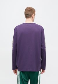 Violets garām piedurknēm T-krekls ar apaļu apkakli, ar dizainu uz kreisās piedurknes. Izgatavots no kokvilnas, tam ir vaļīgs piegriezums un taisna apakšmala.