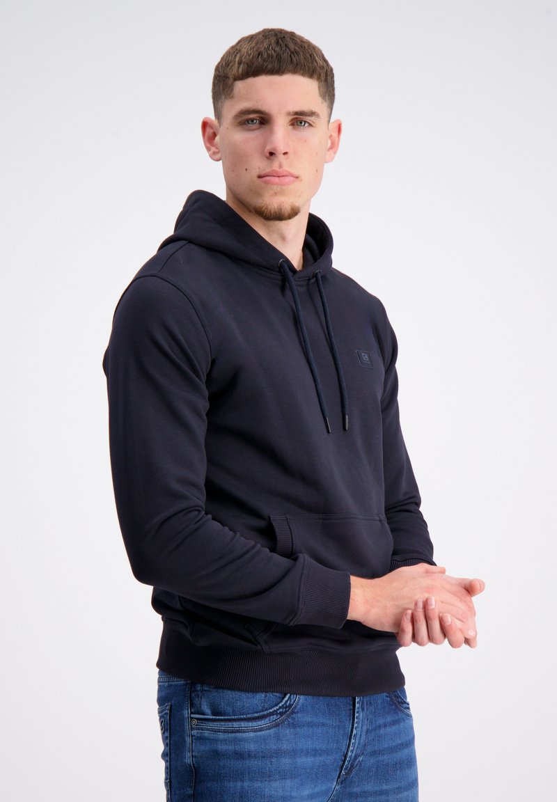 Marineblauer Hoodie mit einer vorderen Känguru-Tasche, Kapuze mit Kordelzug und gerippten Bündchen. Kombiniert mit blauen Jeans, die eine einfache, glatte Stoffoberfläche aufweisen.