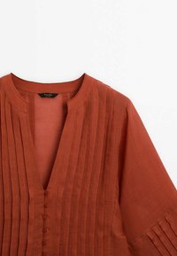 Rustrød bluse med V-hals og knaplukning. Har vertikale plisseringer fra skulder til kant, korte ærmer og en tekstureret finish.