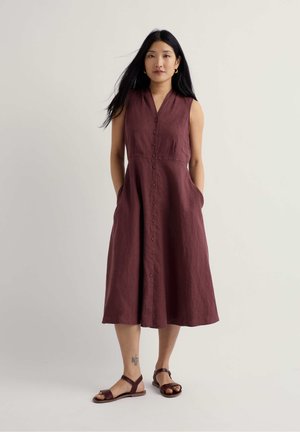 Mujer con cabello largo y negro, vestida con un vestido midi burdeos sin mangas con botones y sandalias a juego, de pie con las manos en los bolsillos.