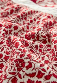 Rød blomsterprint stof med detaljeret design på en cremefarvet baggrund. Teksturen virker blød, med fremtrædende røde blomster og blade.