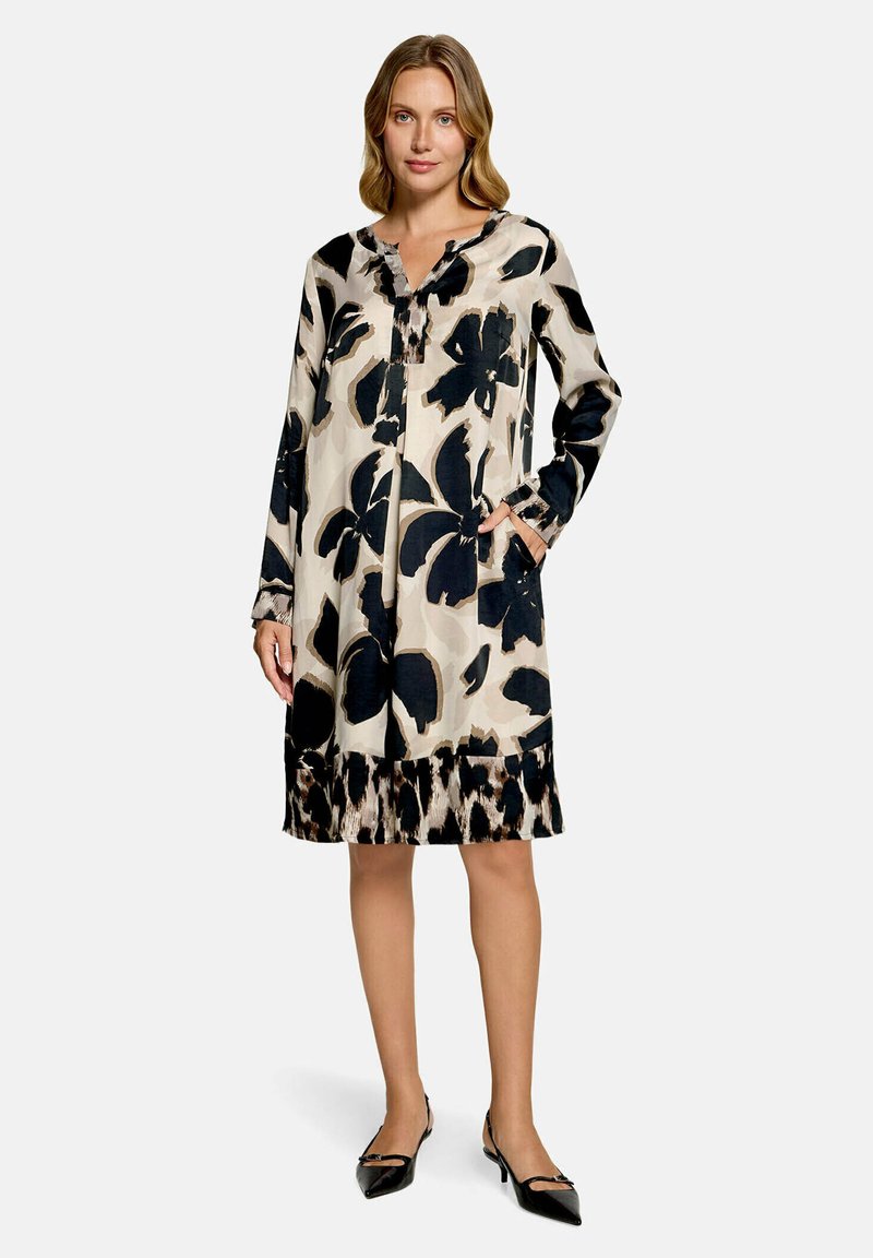 Femme portant une robe beige à manches longues avec de grandes imprimés floraux noirs et des chaussures noires pointues à talons bas, debout devant un fond uni.