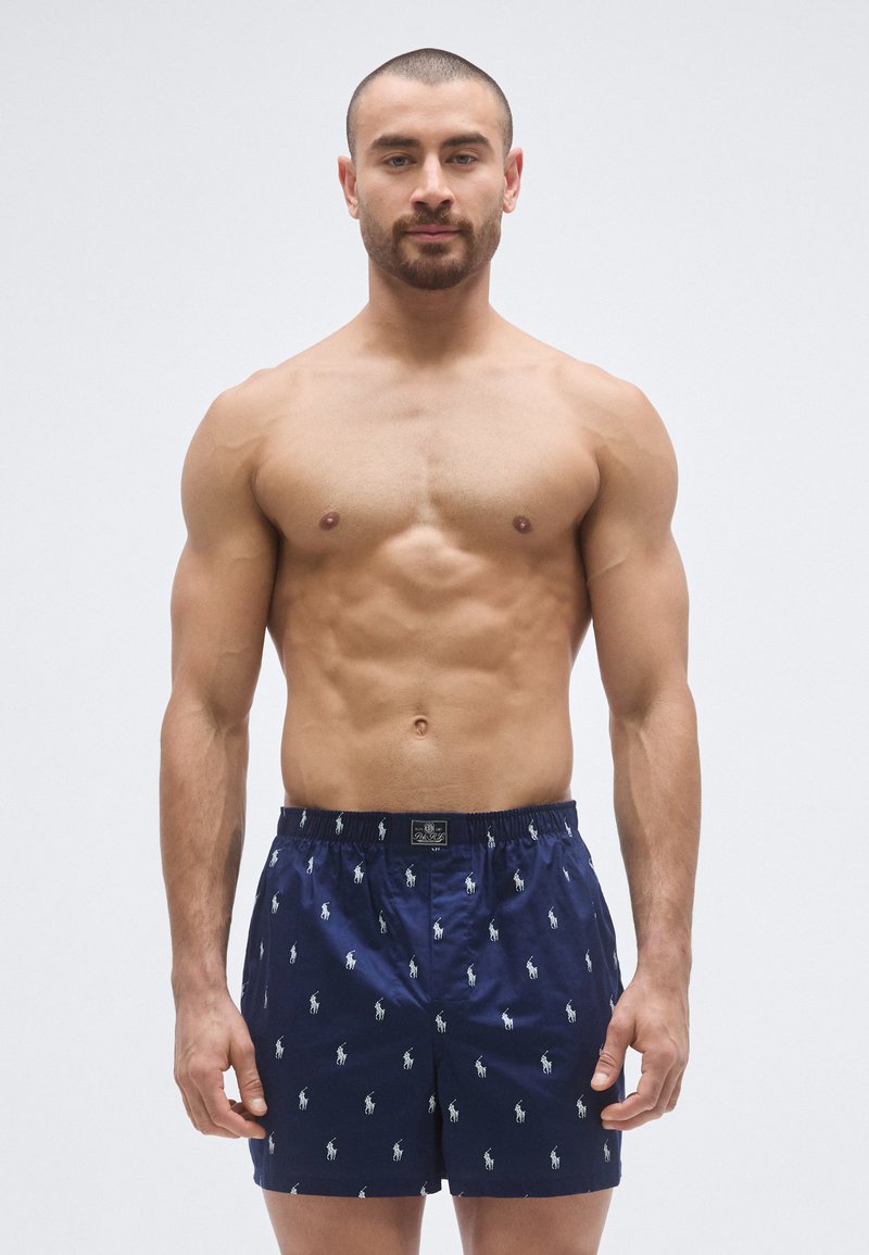 Navy blue swimming shorts with a glossy finish, featuring a repeated white polo logo pattern. Elastic waistband for comfort.