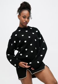 Maglione nero con motivi a cuore bianchi, vestibilità ampia, collo a costine. Abbinato a una gonna nera con orlo decorato da perle.