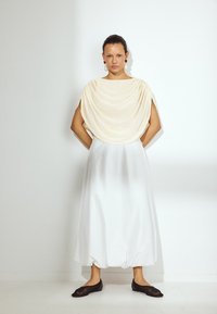 Jasna kremowa bluzka w kształcie narzuty, biała, zwiewna spódnica i ciemne buty wsuwane. Gładkie tekstury materiału, prosty krój, długość do kolan. Minimalistyczny styl.