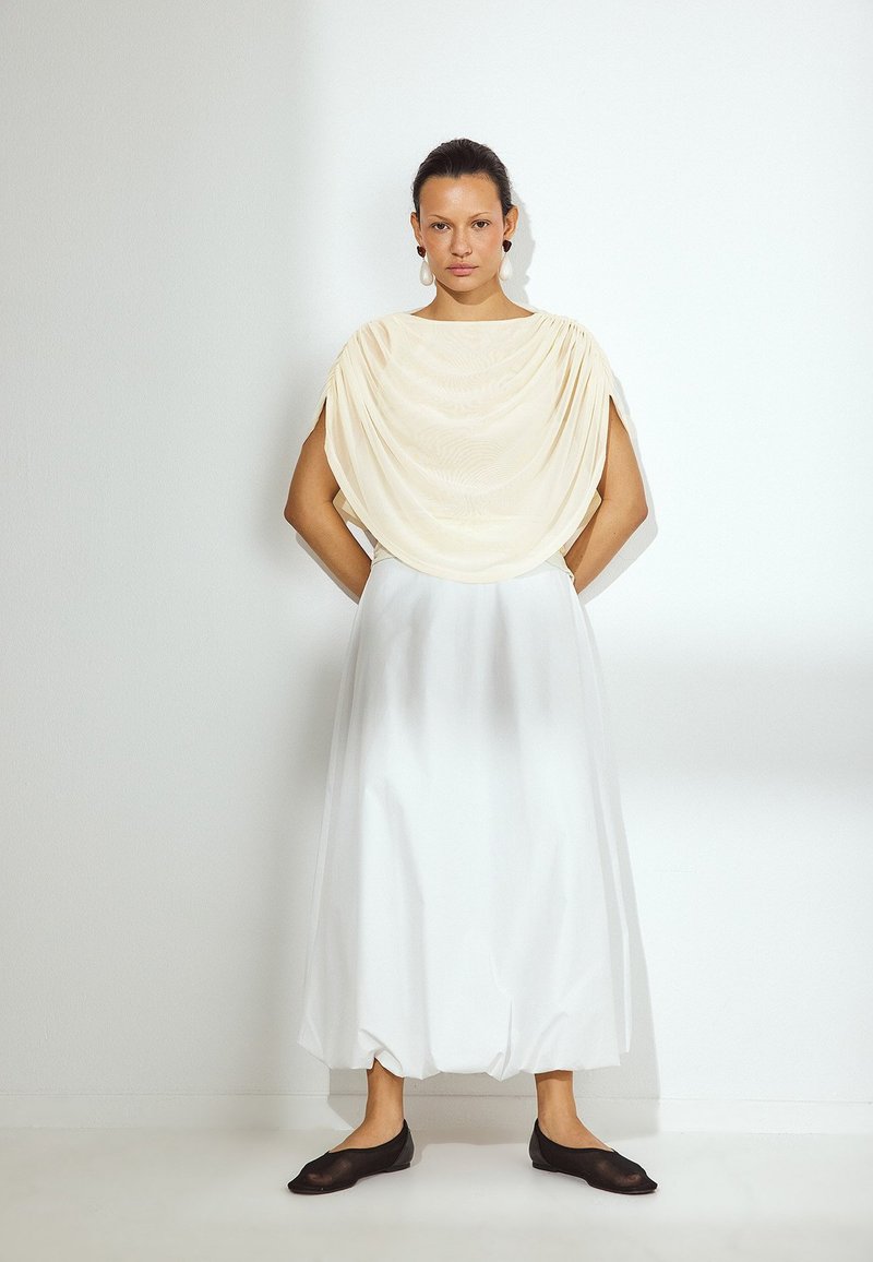 Jasna kremowa bluzka w kształcie narzuty, biała, zwiewna spódnica i ciemne buty wsuwane. Gładkie tekstury materiału, prosty krój, długość do kolan. Minimalistyczny styl.