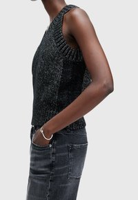 Svart ärmlös stickad topp med texturerat mönster och ribbad halsringning, kombinerad med mörkgrå jeans. Modellen står med ena handen i fickan.