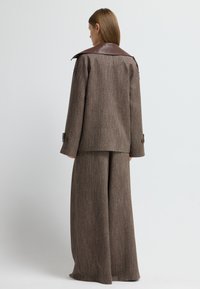 Completo in tessuto marrone con texture, composto da pantaloni a gamba larga e giacca coordinata, con colletto in pelle marrone e polsini con bottoni.
