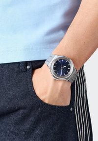 Ezüst óra kék számlappal és rozsdamentes acél hálós pánttal, egy csuklón viselve. Világoskék ing és sötét farmer csíkos részletekkel látható.