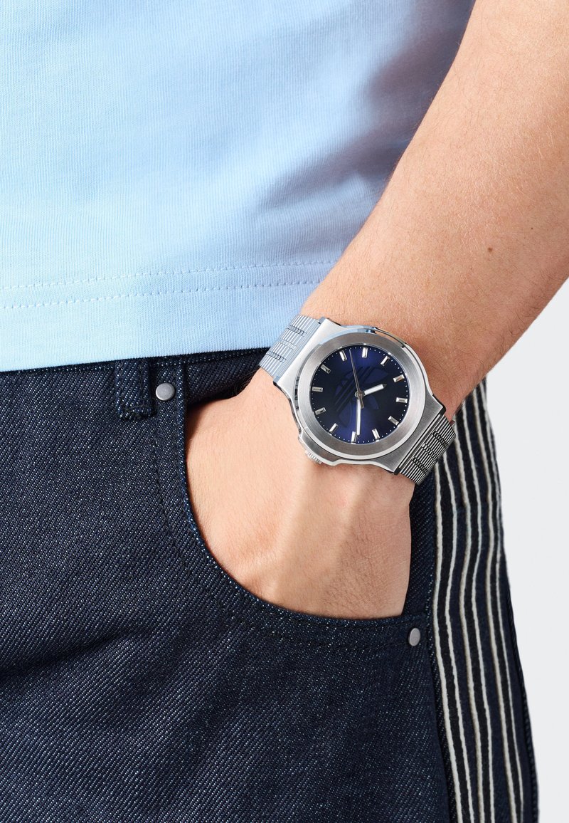 Ezüst óra kék számlappal és rozsdamentes acél hálós pánttal, egy csuklón viselve. Világoskék ing és sötét farmer csíkos részletekkel látható.