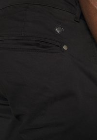 Gros plan d'une poche de pantalon noire avec fermeture éclair, rivet en métal et petit écusson de marque en forme de losange près de la ceinture sur une peau foncée.