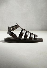 Sandalo in pelle marrone scuro stile gladiatore con suola piatta, cinturino alla caviglia con fibbia e bordi smerlati sulle fasce orizzontali.