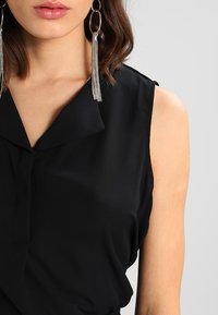Blouse noire sans manches avec col, tissu doux et coupe ample, mise en valeur par de longues boucles d'oreilles à pampilles argentées.