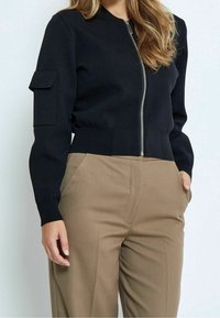 Donna che indossa una giacca nera corta con zip e una tasca sul maniche, insieme a pantaloni beige a vita alta, con le mani nelle tasche.