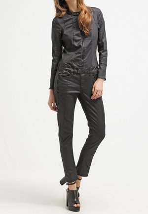 Femme portant une veste et un pantalon moulants en similicuir noir avec des bottines noires à talons ouverts, debout, une jambe croisée.
