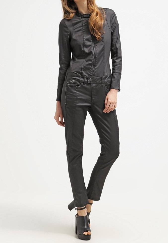 Femme portant une veste et un pantalon moulants en similicuir noir avec des bottines noires à talons ouverts, debout, une jambe croisée.
