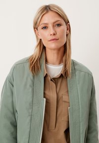 Blonde vrouw met blauwe ogen, gekleed in een groene gewatteerde jas over een bruine blouse en een wit gestreepte onderhemd, staand tegen een effen achtergrond.
