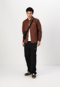 Giacca marrone con bottoni sopra una camicia bianca, pantaloni cargo neri e scarpe da ginnastica nere con lacci bianchi. Presenta un piccolo logo sulla giacca.