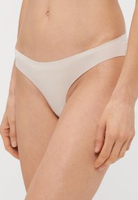 Naga, bezszwowa stringi wykonane z gładkiego, rozciągliwego materiału, o minimalistycznym designie i niskim staniku, z błyszczącym wykończeniem.