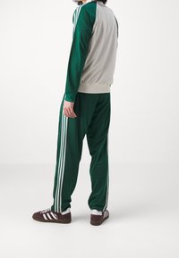 Osoba stojící v zeleno-beige sportovním oblečení s bílými pruhy, v kombinaci s vínovými a bílými teniskami na jednobarevném bílém pozadí.