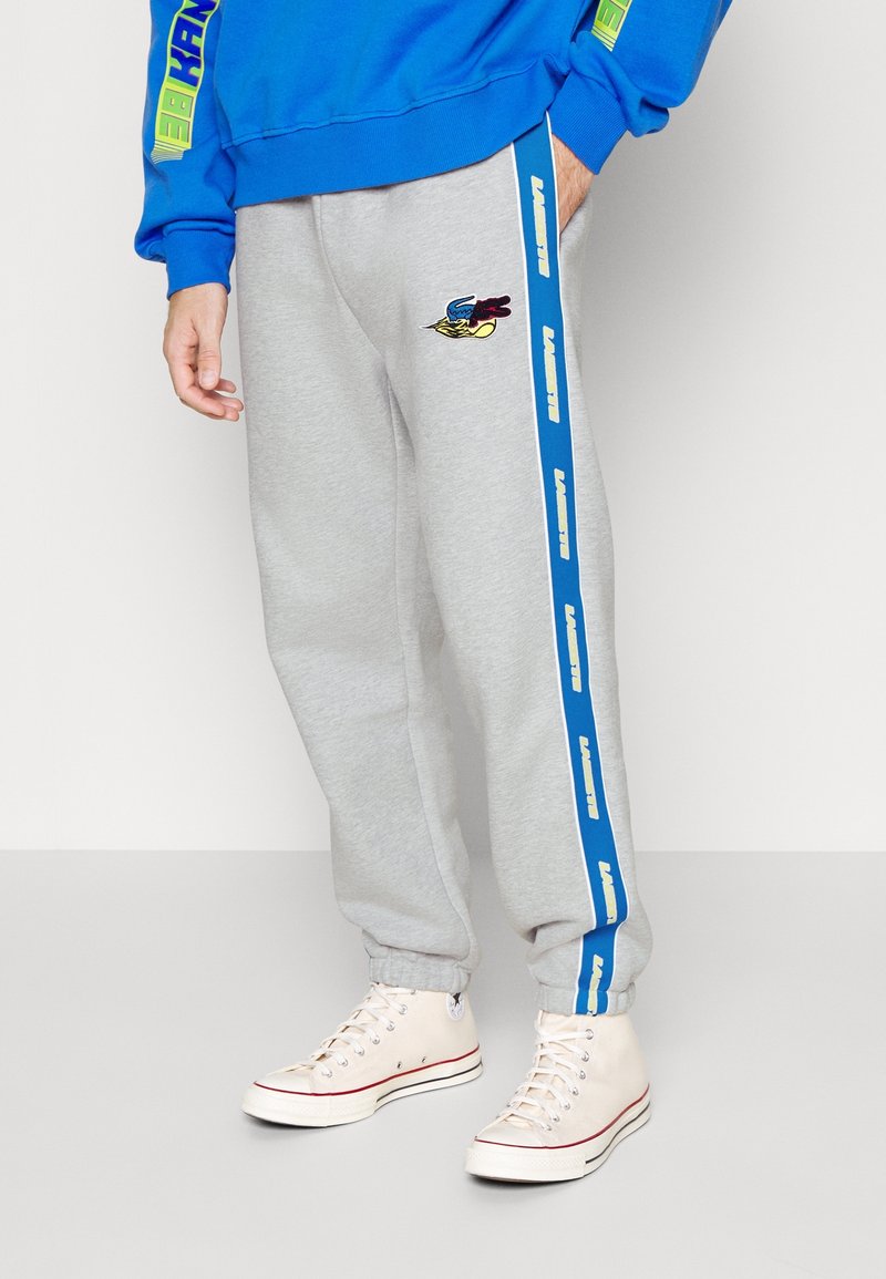 Grå sweatpants med elastiska muddar, med ett blått vertikalt rand med varumärkesnamn och en broderad logotyp. Bärs med ljusa sneakers.