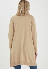Cardigan long beige à manches longues en matériau tricot doux, avec une coupe ample, des poignets côtelés et des fentes sur les côtés pour plus de liberté de mouvement.