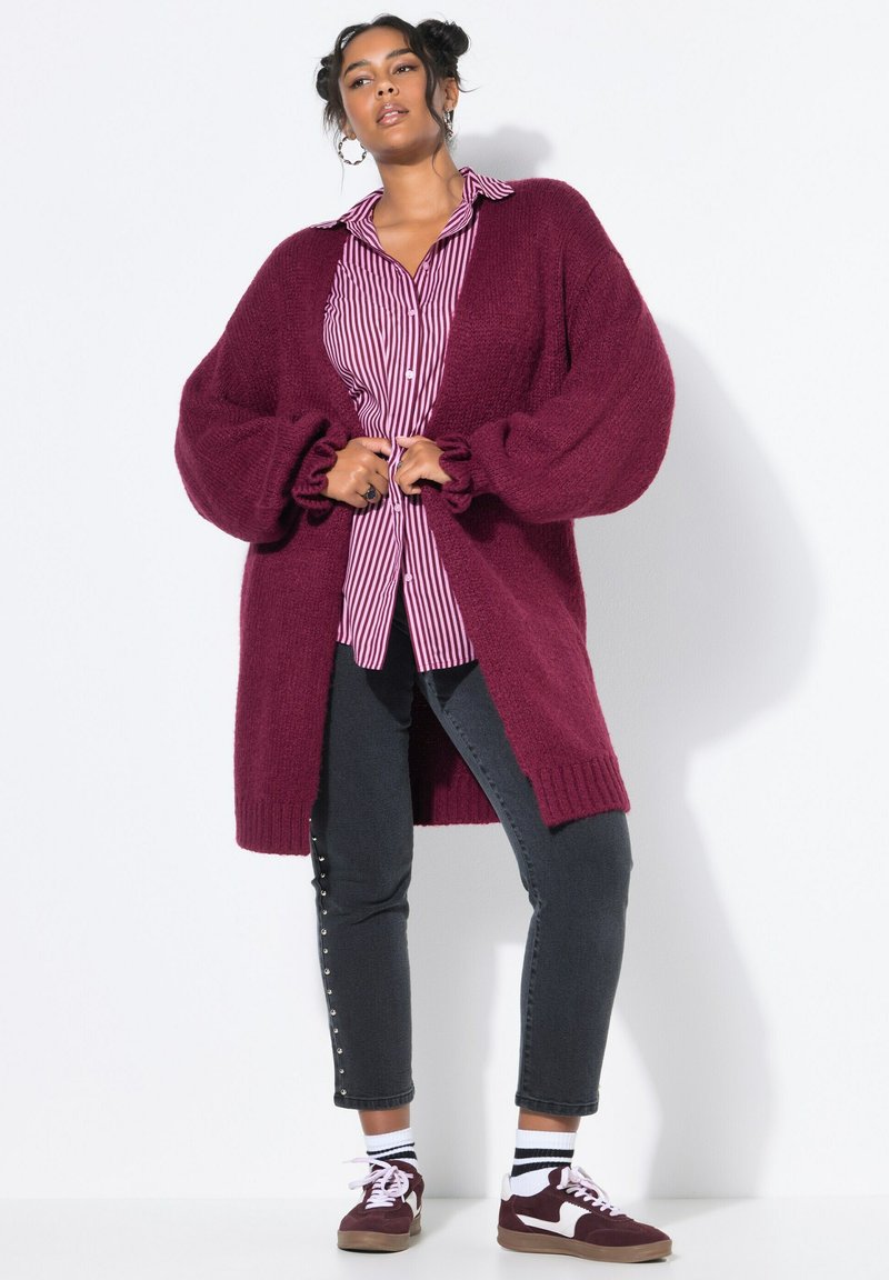 Cardigan en tricot bourgogne avec des manches surdimensionnées, superposé à une chemise boutonnée à rayures roses, associé à un jean noir et des baskets bordeaux.