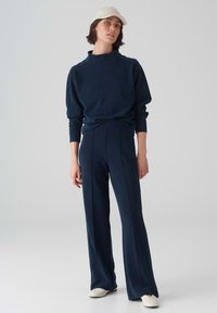 Navyblauwe sweatshirt met hoge hals en verlaagde schouders, gecombineerd met bijpassende op maat gemaakte broek met zijscharen, gedragen met witte schoenen.