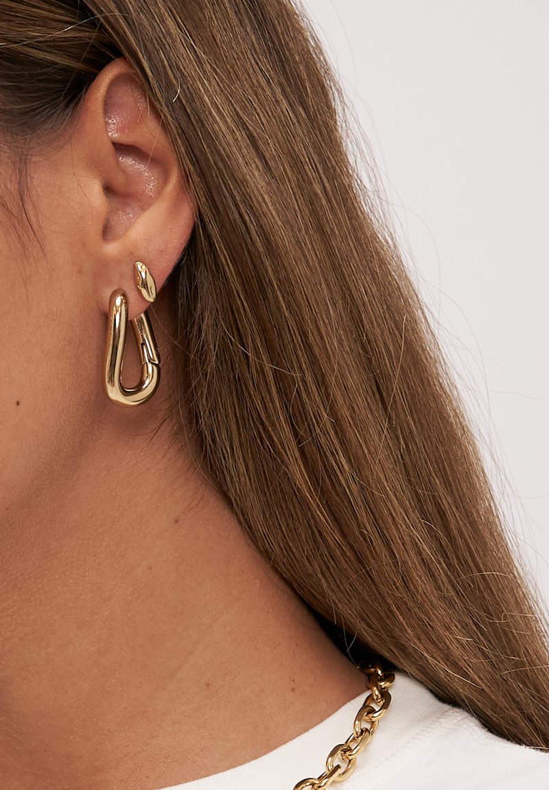 Primo piano di un orecchio femminile con capelli castano chiaro che indossa orecchini a cerchio di forma irregolare in oro e una collana spessa in oro.
