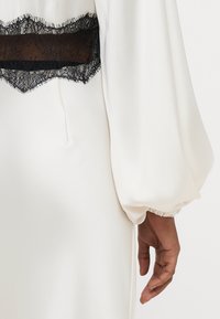 Balta kleita ar caurspīdīgu melnas mežģīnes paneli viduklī. Ar garām pūstām piedurknēm un elastīgām aprocēm. Gluda, teksturēta auduma.