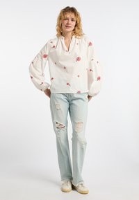Weißes Baumwollbluson mit kurzem Schlitz, verziert mit pinkfarbener Blumenstickerei. Kombiniert mit hellblauen Ripped Jeans und beigen Sneakers.
